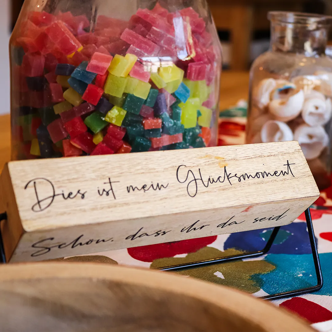 aufsteller aus holz mit einem spruch dies ist mein gluecksmoment im hintergrund verschiede offene glaeser mit inhalt zum basteln