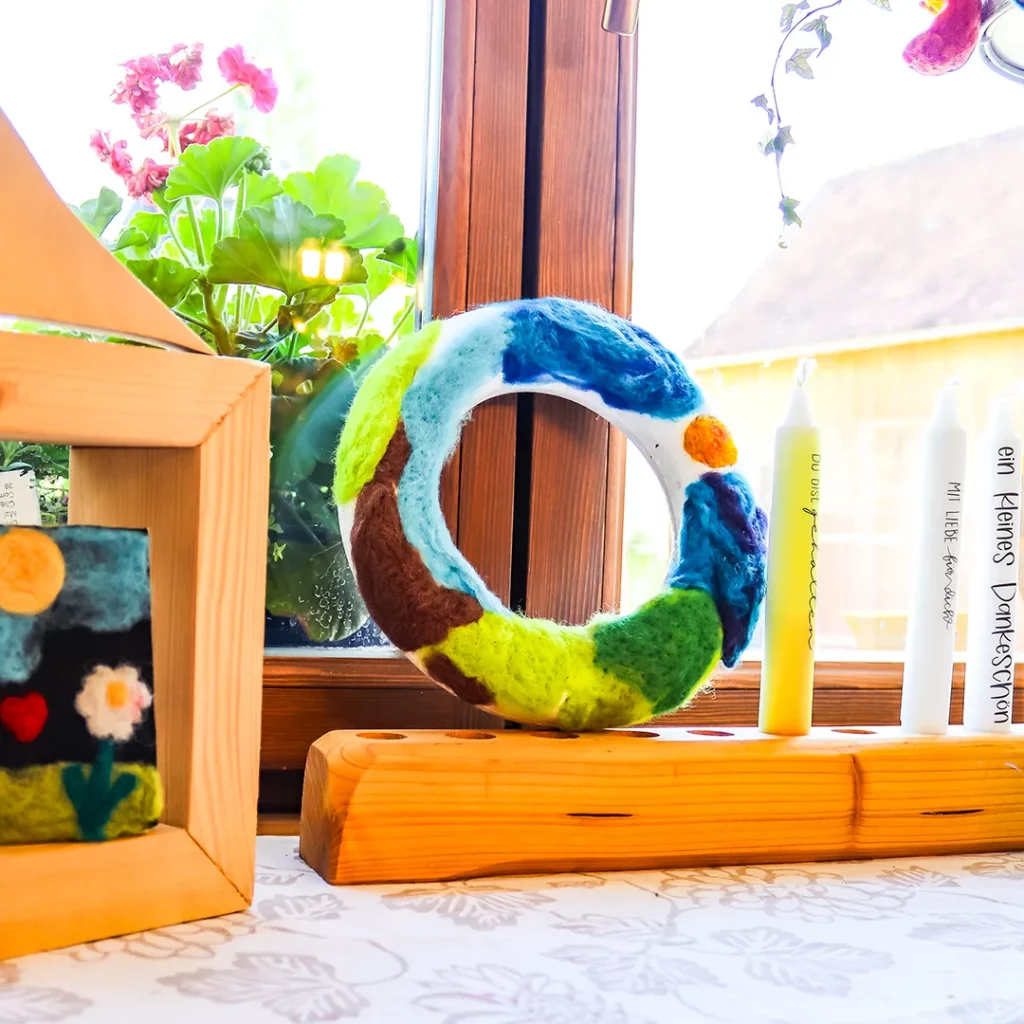 bastel dekoration auf einem fensterbrett mit einem holzrahmen daneben ein ring aus buntem filz mit kerzen auf einem geschliffenen holzkoerper