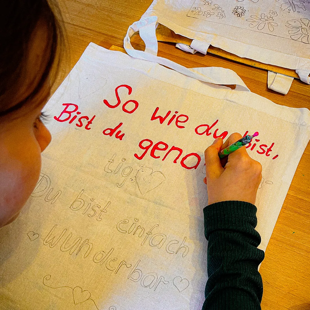 ein maedchen malt mit roter farbe buchstaben aus die zuvor mit bleistift auf eine stofftasche skizziert wurden
