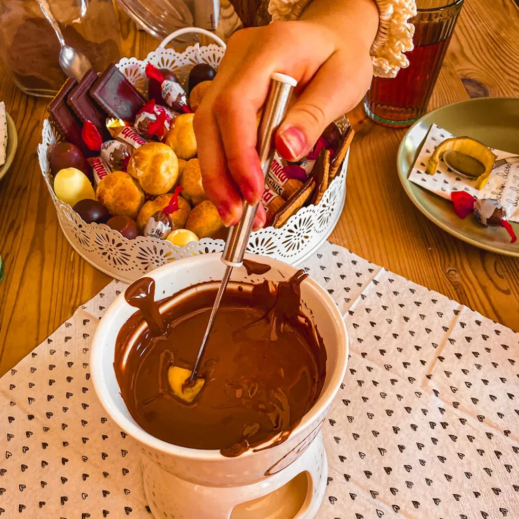mit farbe befleckte hand taucht mit spiesser eine frucht in fluessige schokolade im hintergrund verschiedene suesigkeiten